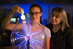 A female student holds a light bulb up to a plasma ball as a female professor looks on. The plasma ball's purple tendrils rise up to the bulb, causing it to illuminate.
