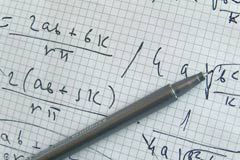 An ink pen lies on top of a piece of graph paper, upon which multiple mathematical equations are scribbled.
