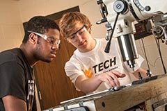 Two students look on their work as they operate a drill press.