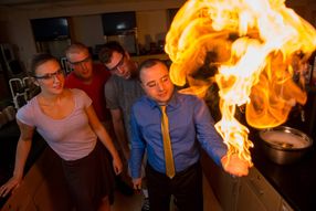 At right, a professor stands in a lab with a massive ball of flame rising from his hand while three students look on in astonishment.